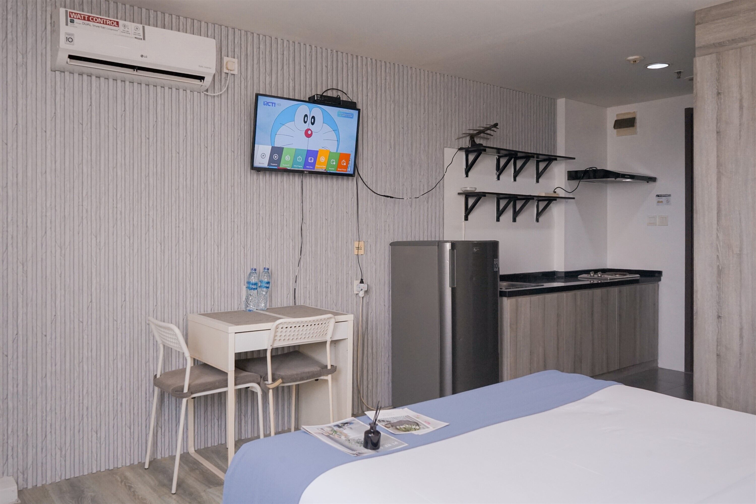 Habitación de hotel con cama, escritorio y silla, televisor montado en la pared, kitchenette con estufa y microondas, y equipo de aire acondicionado.