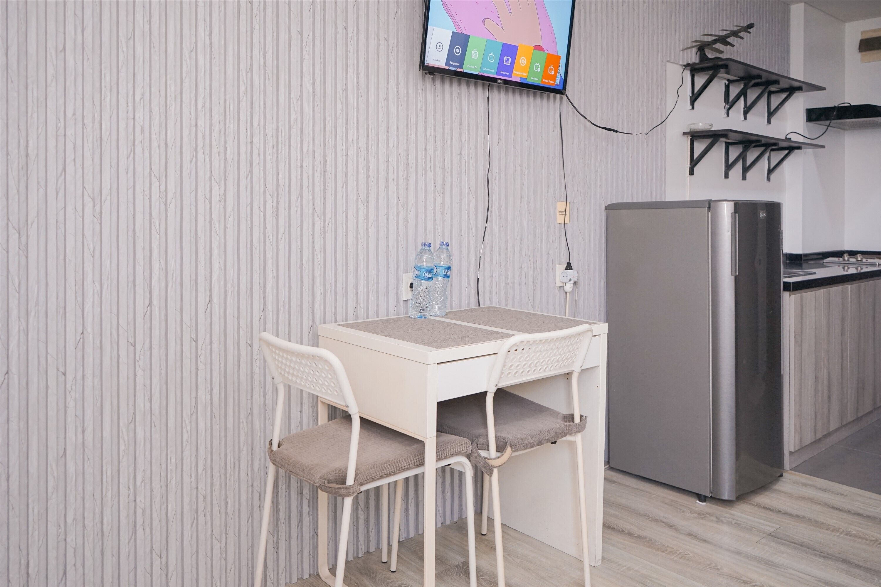 Una kitchenette pequeña con una mesa y sillas blancas, un refrigerador y un televisor de pared que muestra una interfaz colorida.