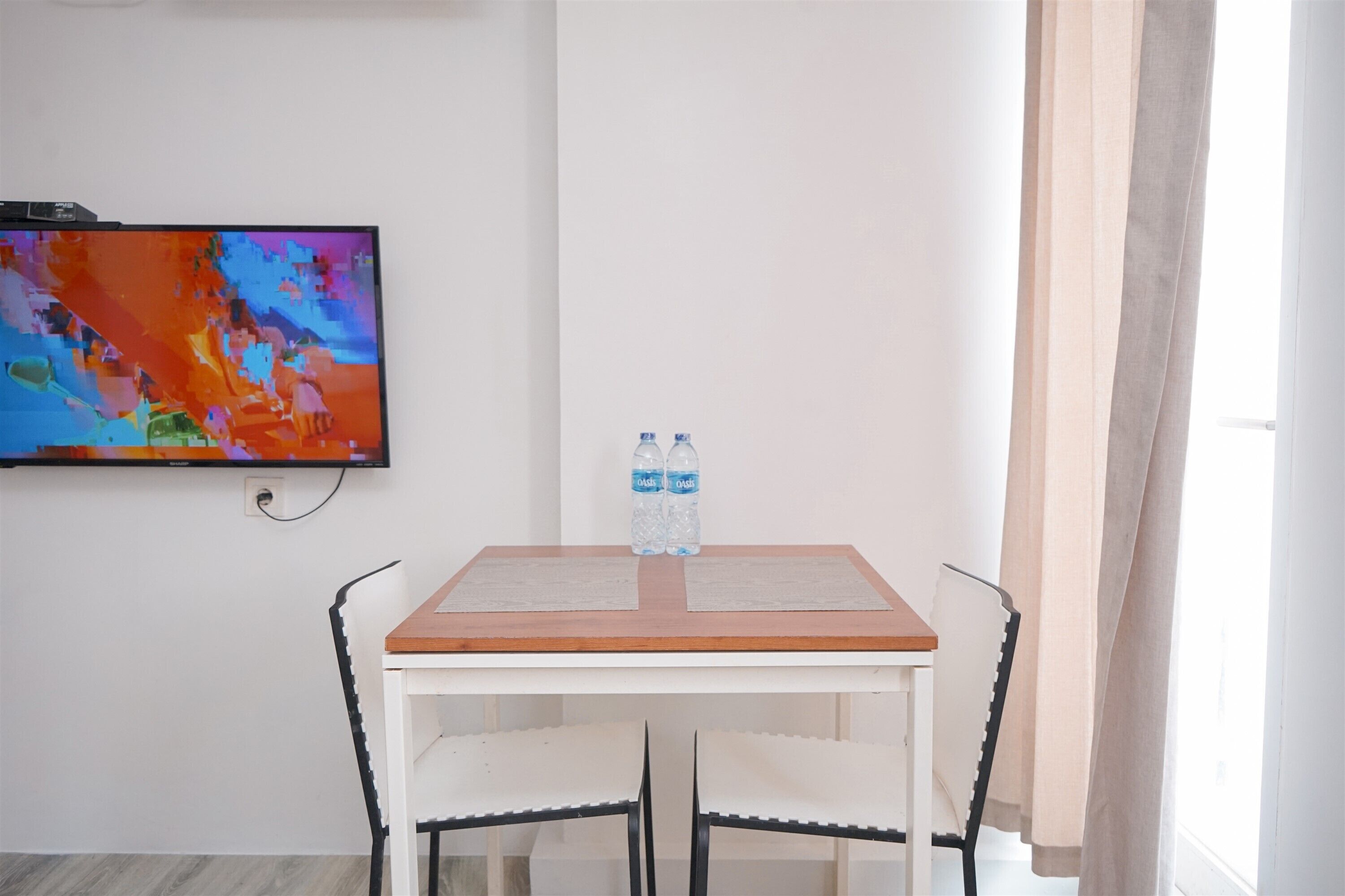 Une petite pièce avec une table blanche et deux chaises, un téléviseur fixé au mur et deux bouteilles d’eau sur la table.