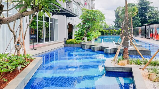 Zona de piscina con suelo de baldosas, rodeada de vegetación y un edificio con grandes ventanales.
