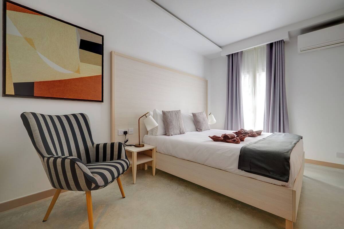 Een moderne hotelkamer met een groot bed, een gestreept fauteuil, een nachtkastje met lampen en een groot abstract schilderij aan de muur.