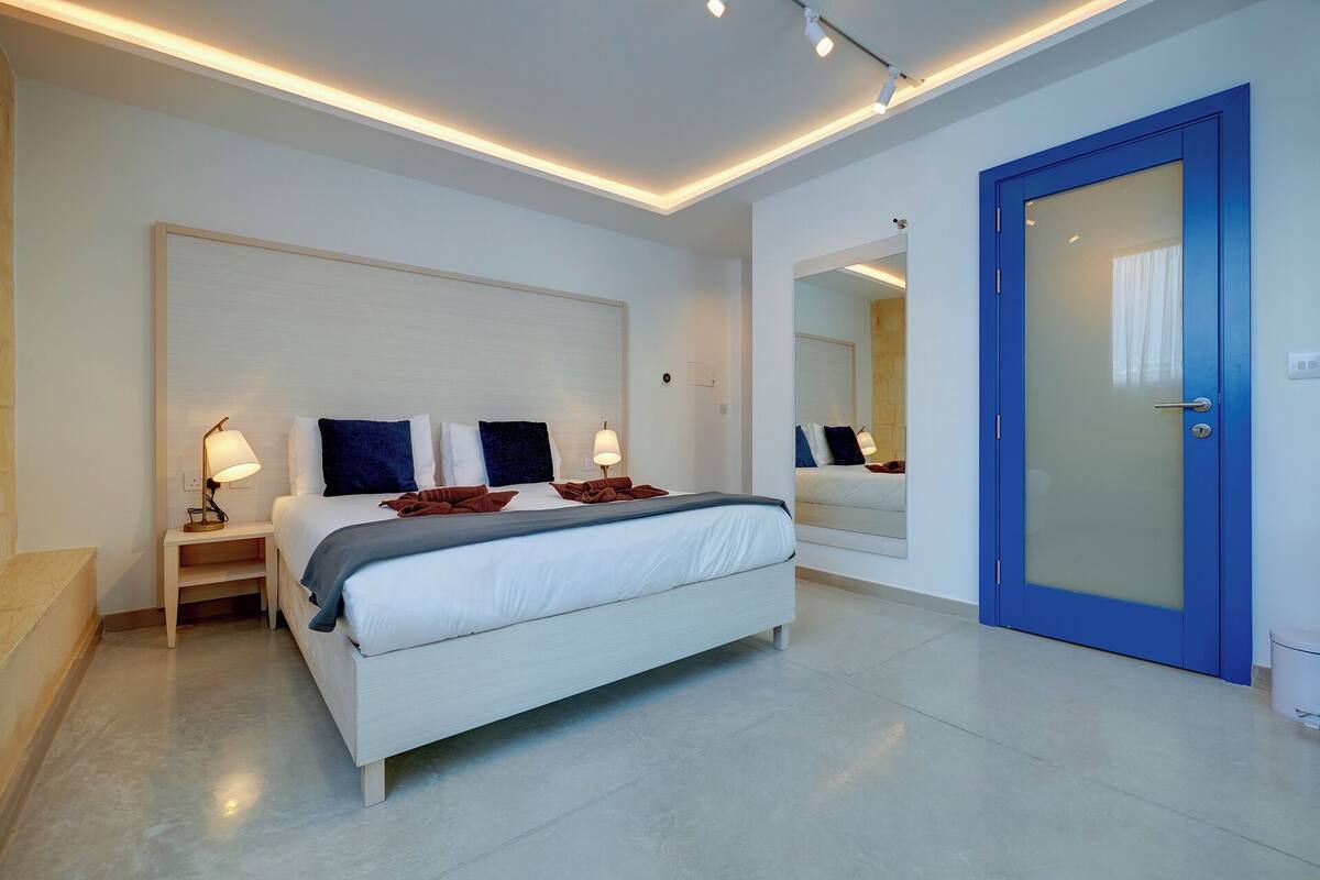 Een moderne slaapkamer met een bed, nachtlampjes, een grote spiegel en een blauwe deur.
