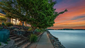 A waterfront pathway with a stone wall and a large tree on the left, leading to a house with a well-lit exterior.