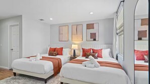 A hotel room with two beds, white bedding, red and brown pillows, a white rug, and artwork on the walls.