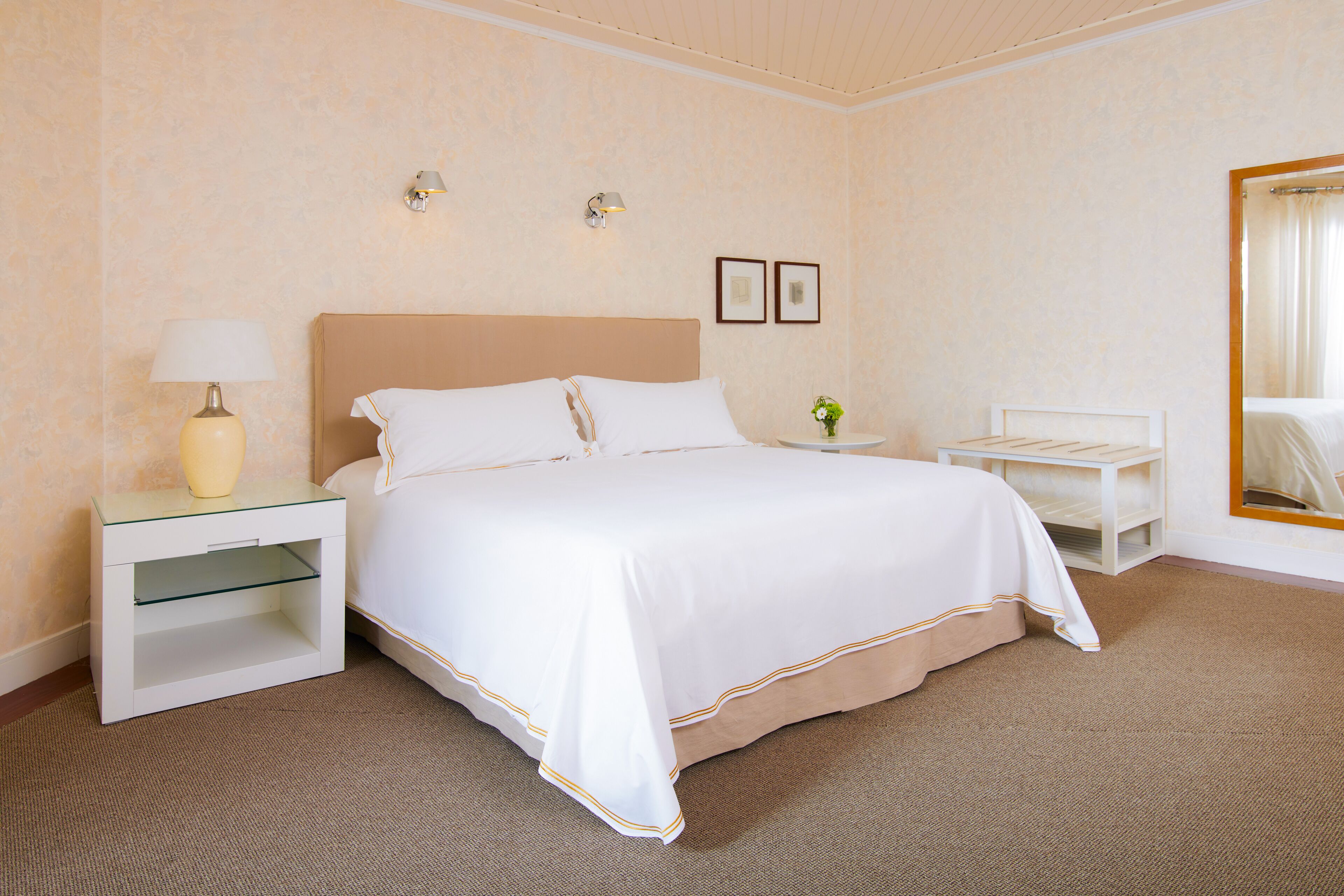 A hotel room with a large bed, a bedside table with a lamp, a small side table with a vase, and a mirror on the wall.