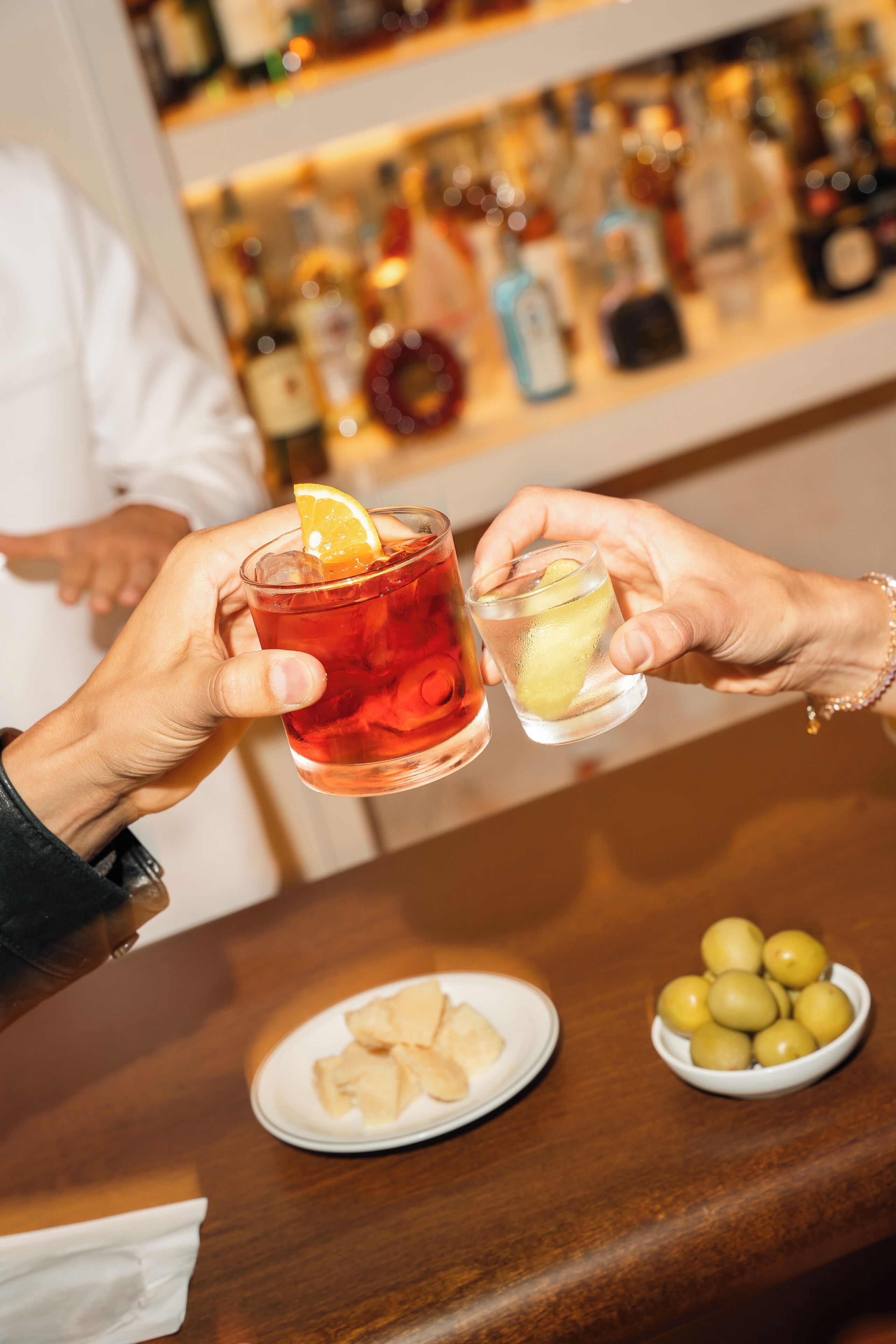 Two hands holding glasses of a red beverage with a lemon slice, a plate of crackers, and a bowl of olives on a wooden bar counter.