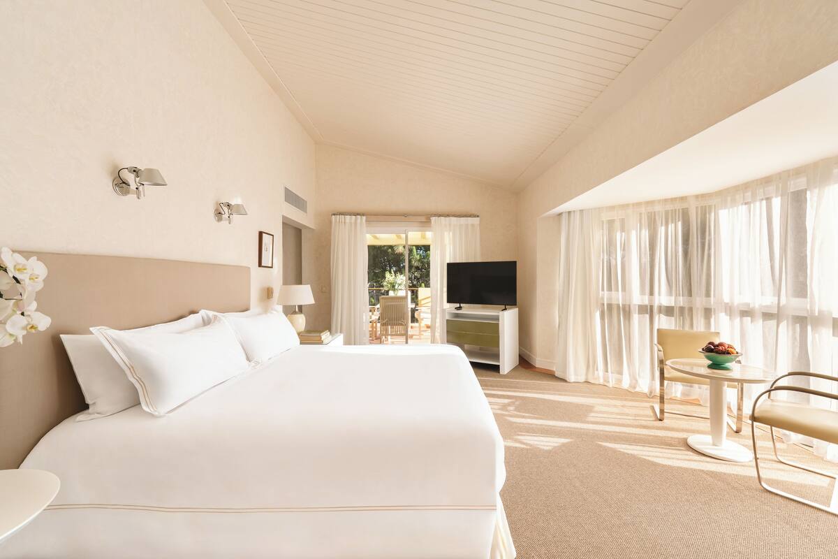 Ein Hotelzimmer mit einem großen Bett, einem Fernseher, einem Essbereich mit Tisch und Stühlen sowie einem Balkon mit Vorhängen.
