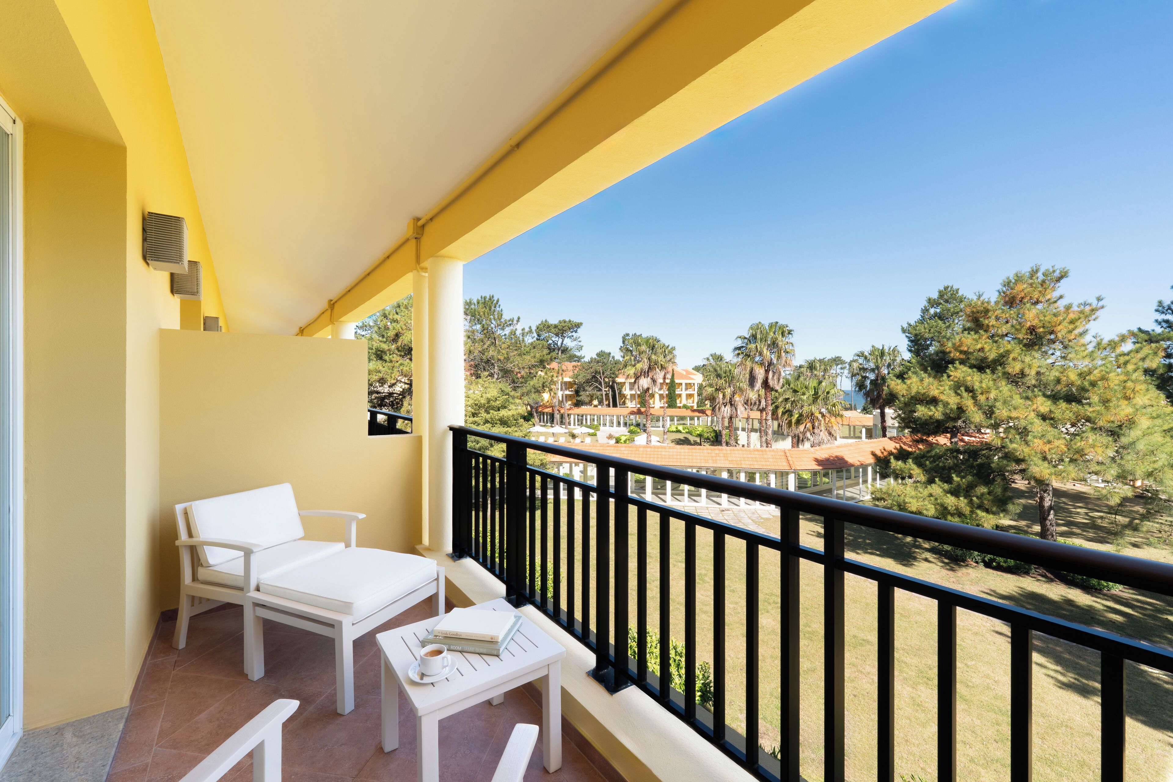 A balcony with a white table and chairs, a small table with a book and a cup, and a view of a residential area with trees and buildings.
