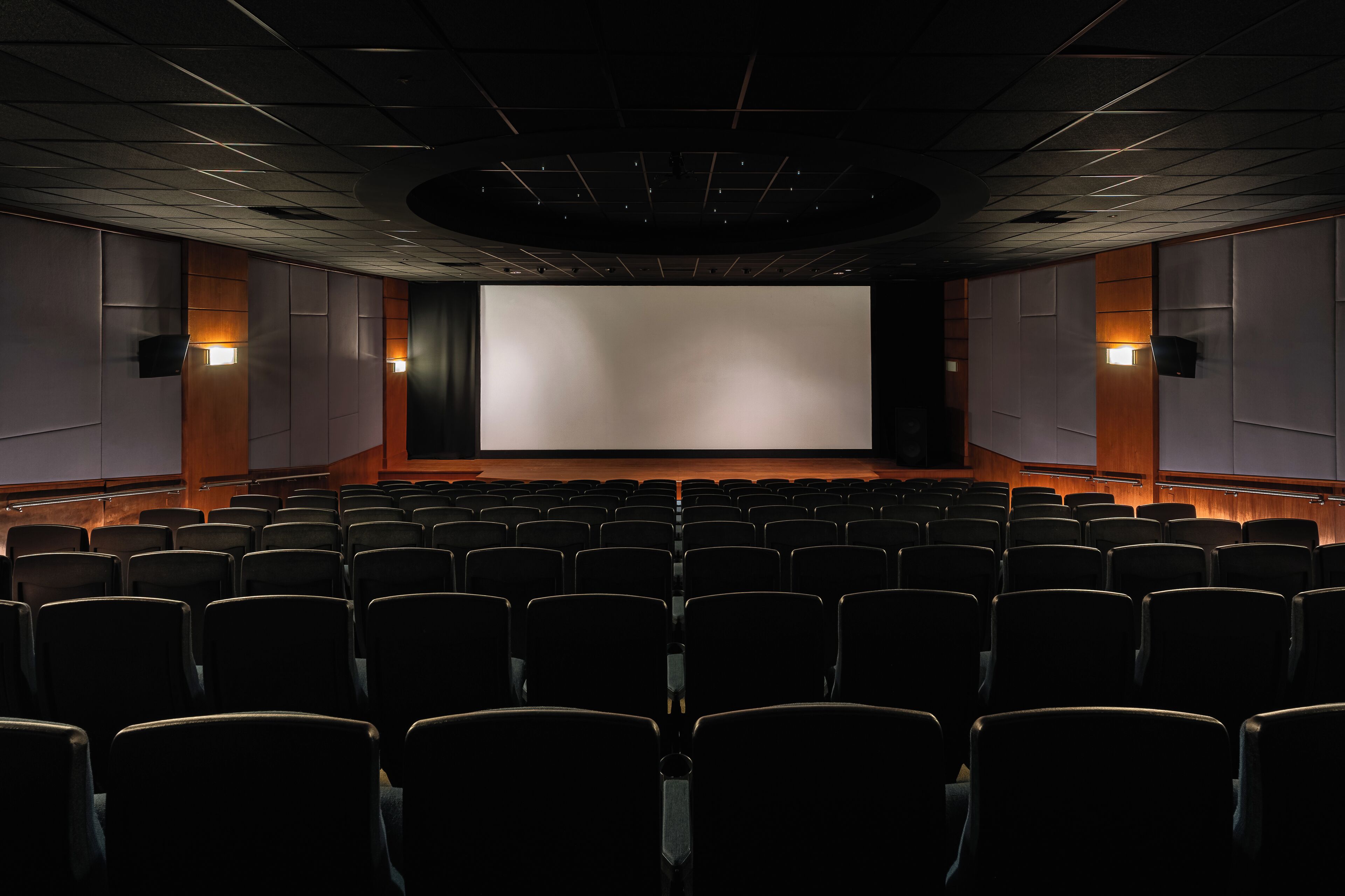 A movie theater with rows of seats, a large screen, and wall-mounted speakers.