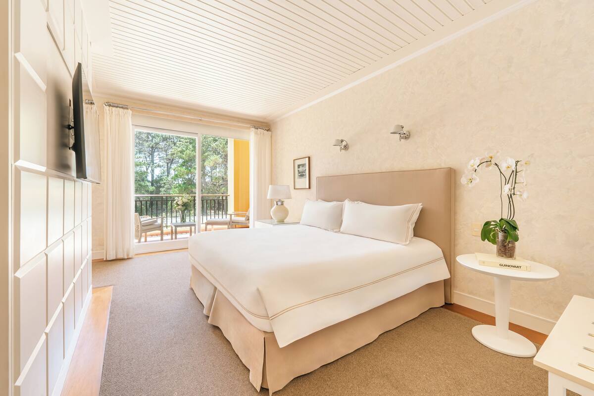 Ein geräumiges Schlafzimmer mit einem großen Bett, einem Nachttisch, einem an der Wand montierten Fernseher und einem Balkon mit einem Sessel und einer Pflanze.