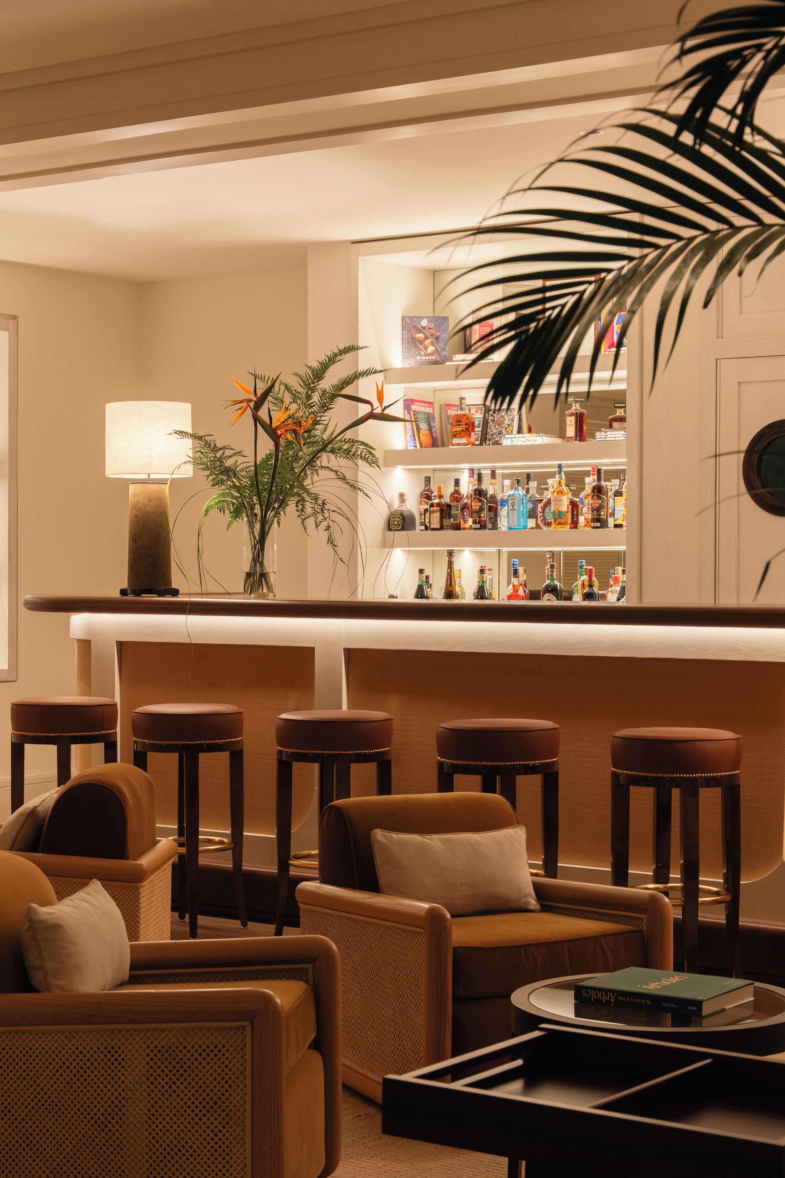 A bar area with a wooden counter, bar stools, and a variety of liquor bottles. There is a potted plant with orange flowers and a lamp on the counter.