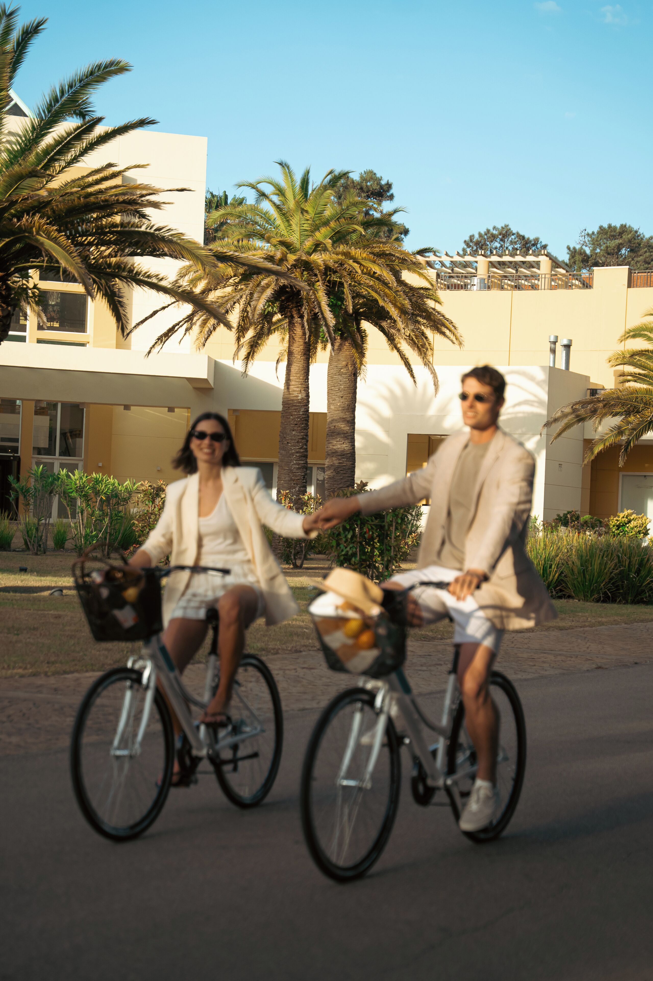 Deux personnes à vélo, dans un cadre extérieur, avec des palmiers et un bâtiment moderne en arrière-plan.
