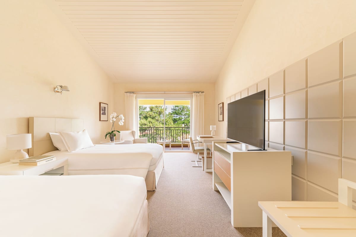 Ein Hotelzimmer mit zwei Betten, einem Schreibtisch, einem Fernseher und einem Balkon mit Blick auf Grünflächen.