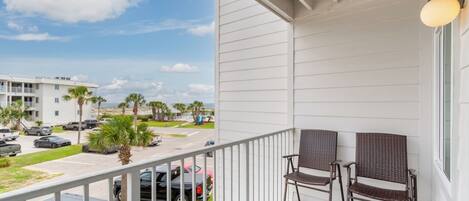 A balcony with two wicker chairs, a railing, and a view of a parking lot and buildings.