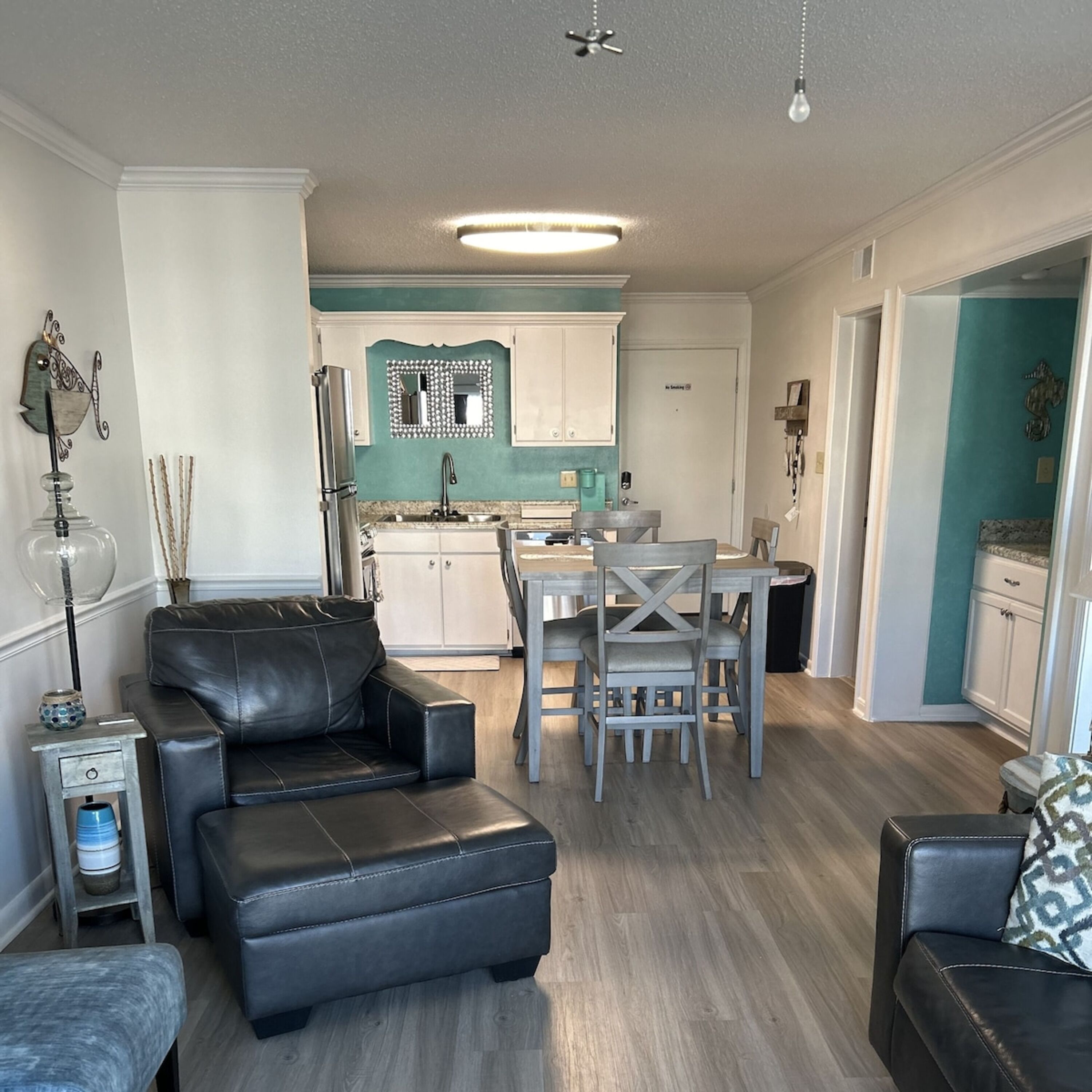 Un salon moderne avec un canapé en cuir noir, un coin repas avec une table et des chaises en bois, et une cuisine avec des meubles blancs et un dosseret de couleur turquoise.