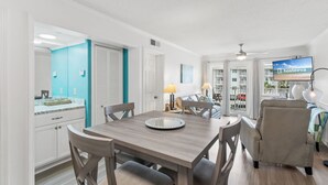 A dining area with a wooden table and chairs, a television mounted on the wall, and a view of the outside through a window. - Plantation 2215 (Gulf Shores)