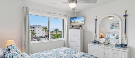A bedroom with a bed, a television, a dresser, and a window with a view of palm trees and buildings.