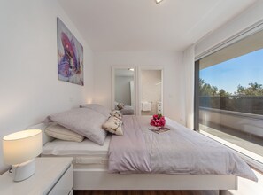 Ein modernes Schlafzimmer mit einem großen Bett, einer Nachttischlampe, einem Spiegel und Blick ins Freie.