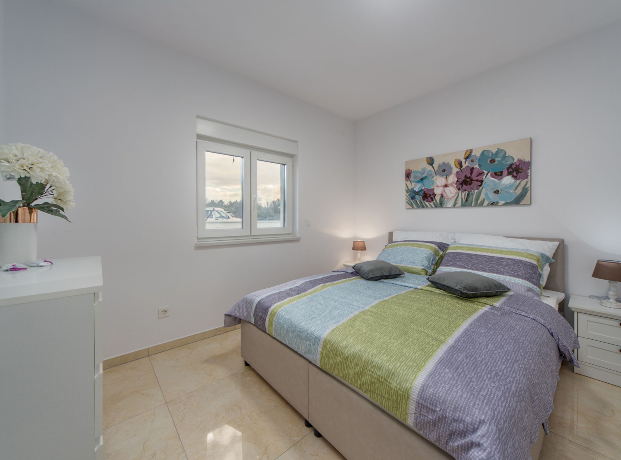 A bedroom with a bed, a window, a bedside table, a lamp, and a painting on the wall.