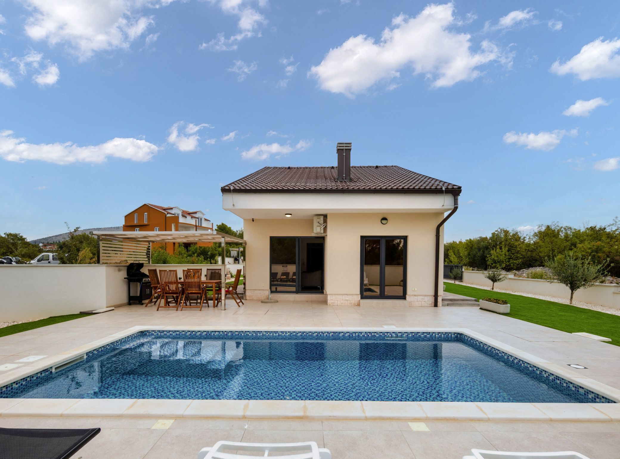 Et moderne hus med swimmingpool, en terrasse med udendørs møbler og en klar blå himmel.