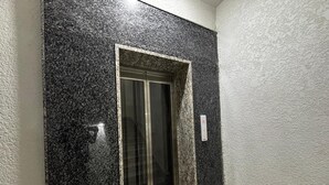 A narrow hallway with a glass elevator door and a textured wall. - Aster Studio Serviced Apartment (Gurugram)