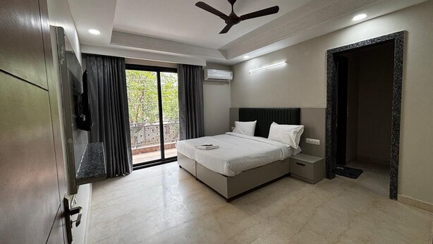 A modern bedroom with a bed, bedside tables, a ceiling fan, and a large window with curtains. - Aster Studio Serviced Apartment (Gurugram)