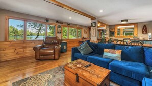 Un salón amplio con un sofá azul grande, un sillón de cuero marrón y una mesa de centro de madera. Amplias ventanas ofrecen vistas a un lago y árboles.