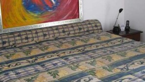 Una habitación con una cama, una colcha estampada, un cuadro abstracto y colorido en la pared, y un escritorio con una lámpara y un teléfono.
