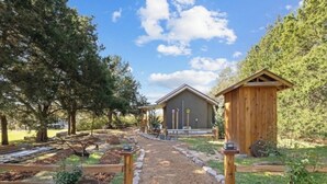 A rustic cabin with a gravel path leading to it, surrounded by trees and garden beds.