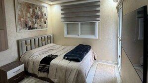 Un dormitorio con cama, una mesita de noche, televisor montado en la pared, ventana con persianas y unidad de aire acondicionado instalada en el techo.