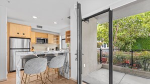 A modern kitchen with a marble countertop, stainless steel appliances, and wooden cabinets. There are two bar stools and a sliding glass door leading to a balcony with a view of a garden and a fence.