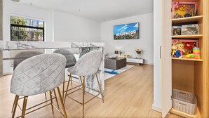 A modern kitchen with marble countertops, a dining area with grey upholstered chairs, a TV displaying 'Hollywood', and a wooden cabinet with toys.