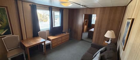 Una habitación con piso de madera, un sofá marrón, una mesa de madera con sillas, una televisión, un ventilador de techo y una ventana con cortinas.