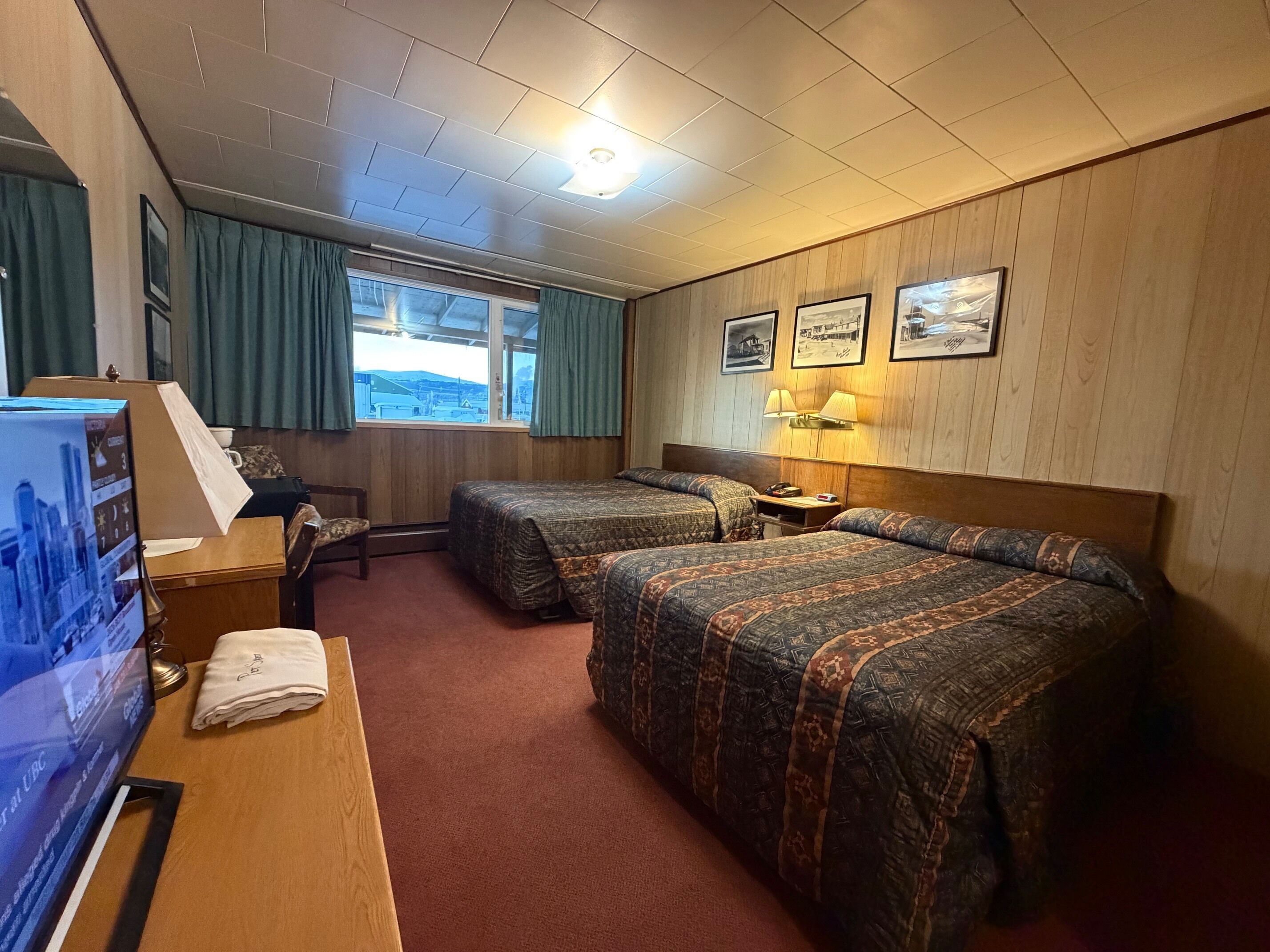 Habitación de hotel con dos camas, un escritorio con computadora, una ventana con cortinas y cuadros en la pared.