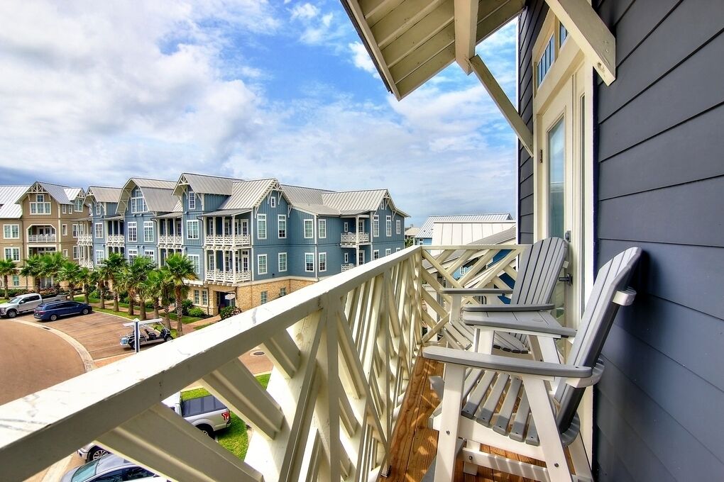 A balcony with white railings and two Adirondack chairs, overlooking a row of townhouses with gabled roofs and a parking lot in the background.