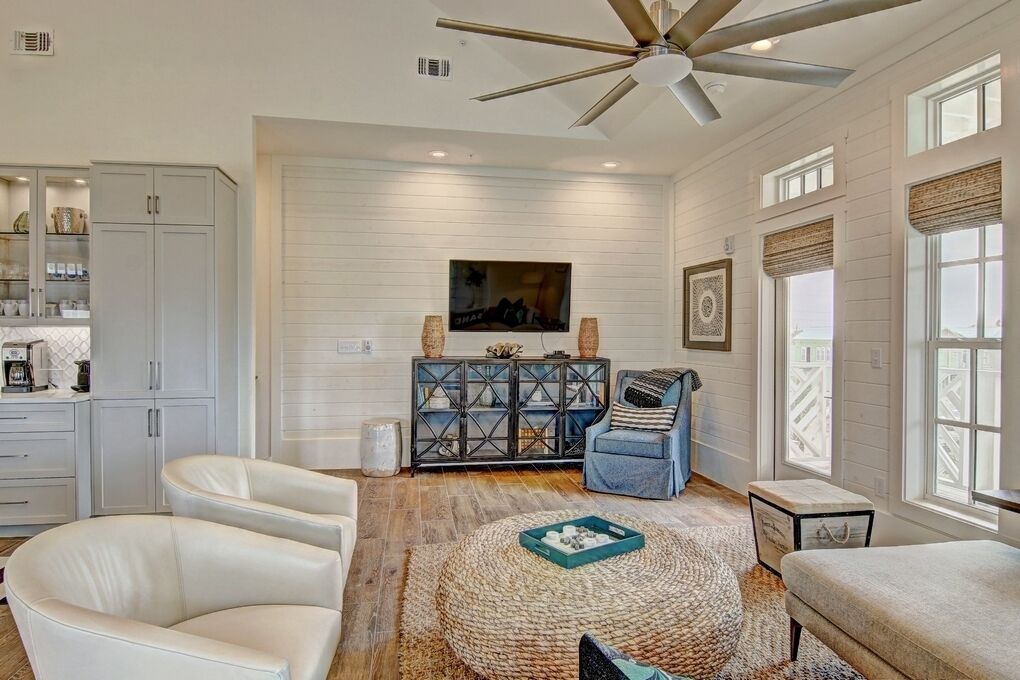 A spacious living room with a ceiling fan, a television mounted on the wall, and a large window with blinds.