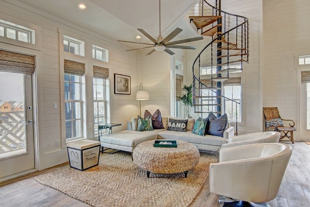 A spacious living room with a large window, a ceiling fan, a spiral staircase, and a sofa with decorative pillows.