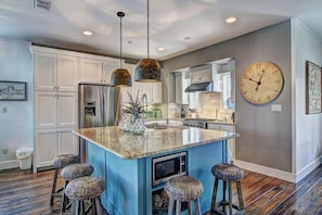 A modern kitchen with a central island, white cabinetry, stainless steel appliances, and a large wall clock. - Shore Beats Workin' 216 BE (Port Aransas)