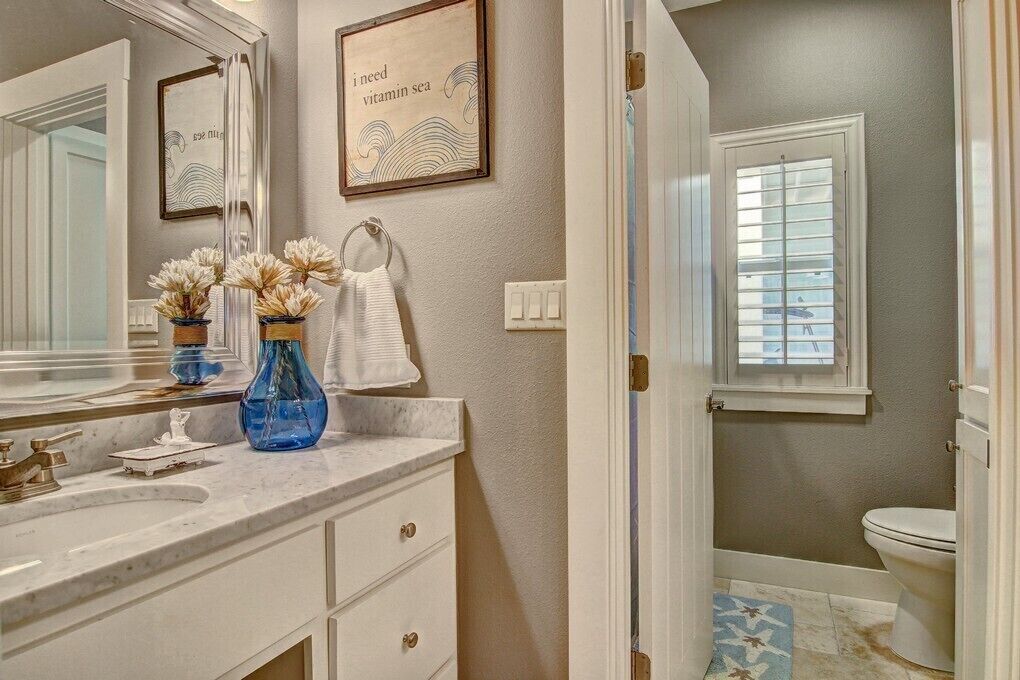 A bathroom with a white vanity, a large mirror, two vases with dried flowers, a towel, and a framed quote on the wall.