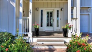 Une maison blanche avec un porche, le numéro « 216 » sur le mur, et deux grands pots de fleurs ornés de plantes.