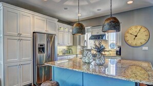Une cuisine moderne avec des meubles blancs, un réfrigérateur en acier inoxydable, un îlot central en granit avec des tabourets et une grande horloge murale.