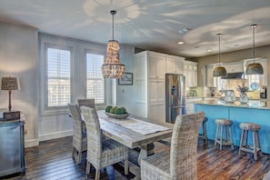 A dining area with a wooden table and wicker chairs, a chandelier, and a kitchen with white cabinets and a blue island. - Shore Beats Workin' 216 BE (Port Aransas)