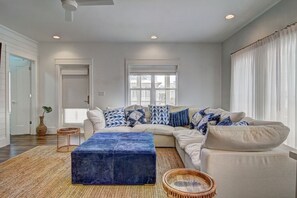 A modern living room with a sectional sofa, a blue ottoman, and a large window with curtains. - Shore Perfection 118 CE (Port Aransas)