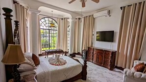 A spacious bedroom with a large bed, a ceiling fan, a TV, and a view of greenery through a window. - Villa San Juan a special place in Tela (El Triunfo de La Cruz)