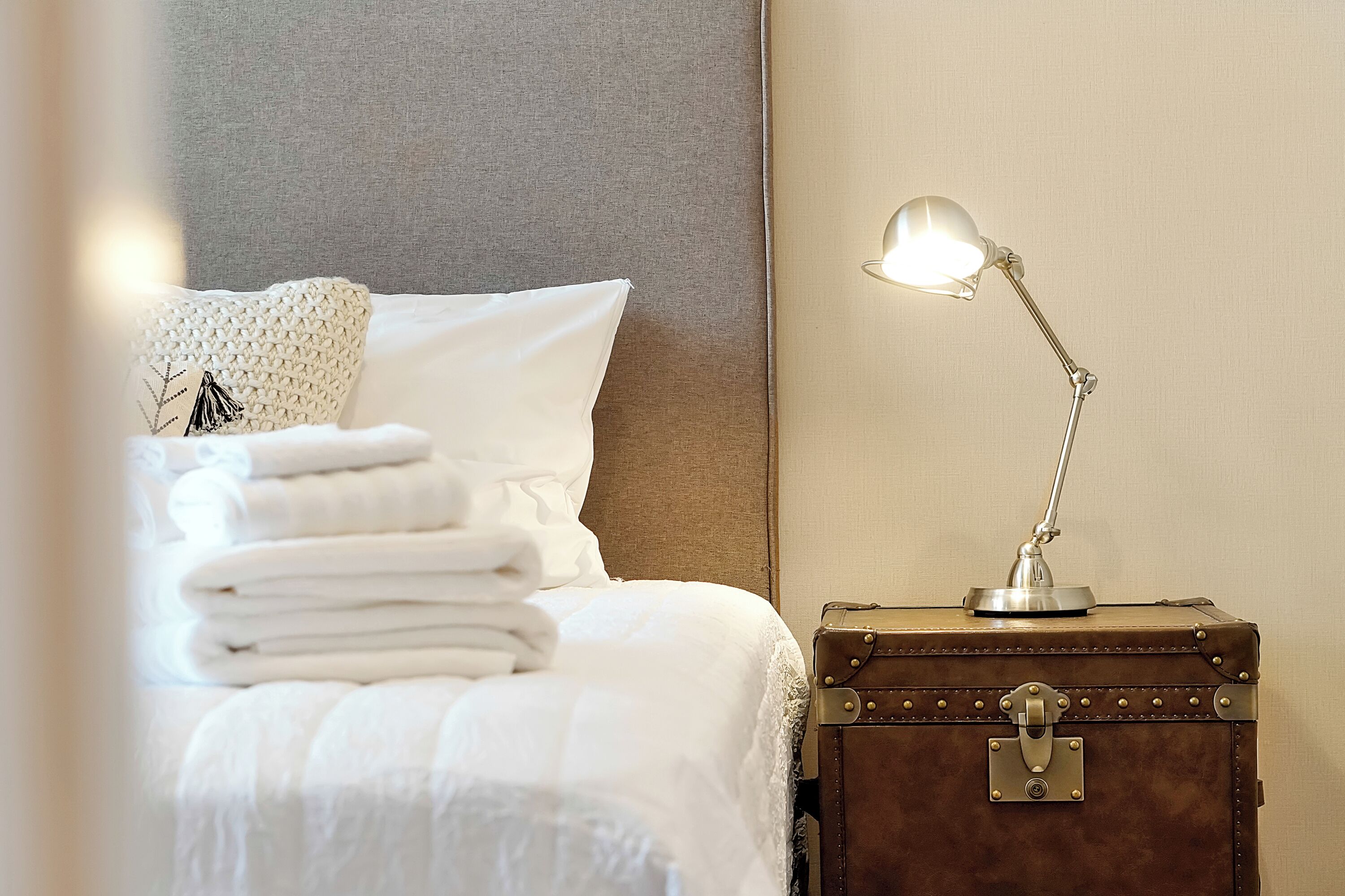 Een net opgemaakt bed met opgevouwen witte handdoeken, een bedlamp op een nachtkastje in vintage-stijl, en een hoofdbord tegen een effen muur.
