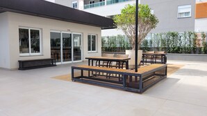 An outdoor patio area with a seating arrangement, a large potted tree, and a building with large windows and a balcony. - Comfortable apartment for up to 3 people near USP - Shopping - Aeroclube (Jardim Brasil Bauru)