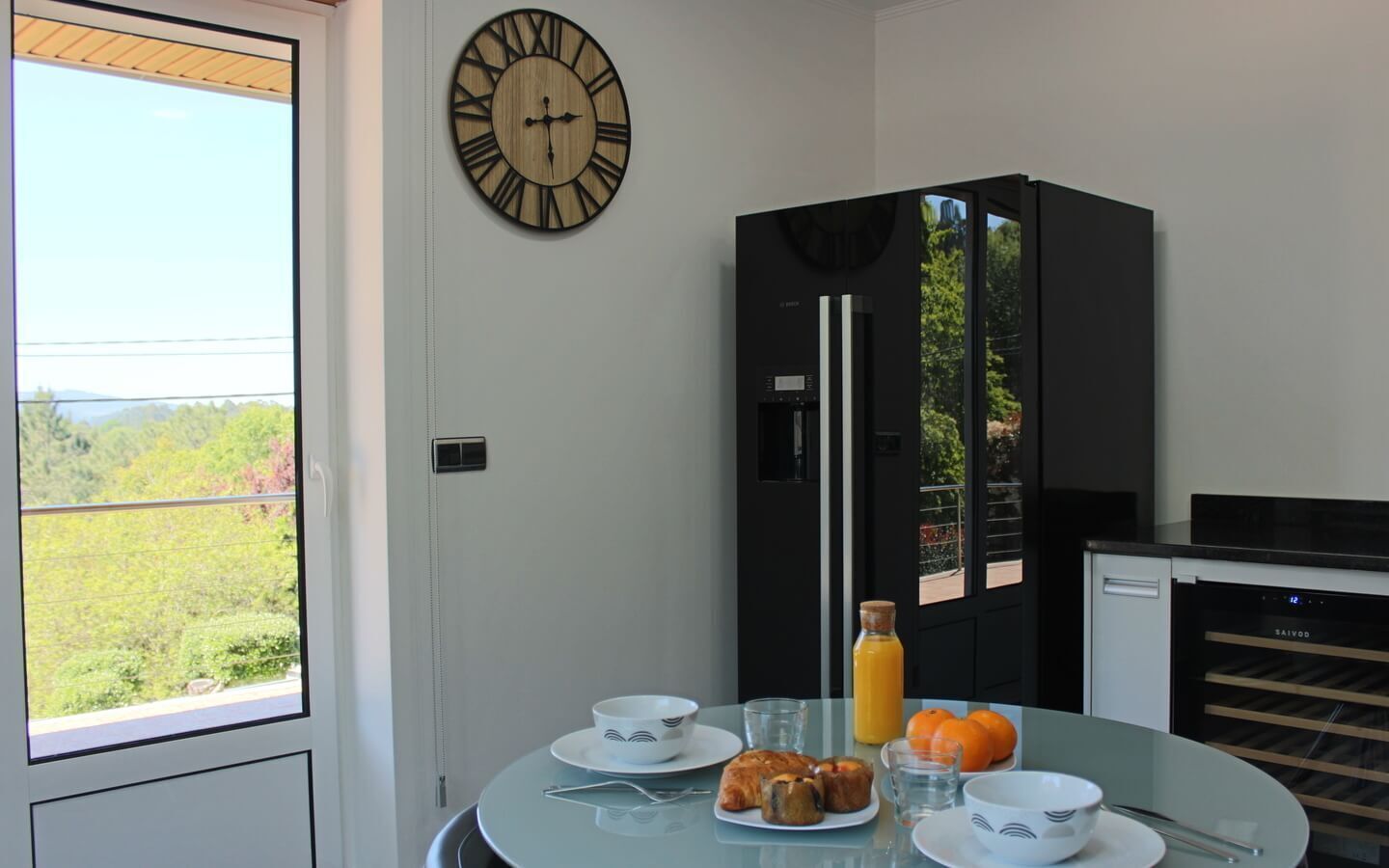 Una cocina moderna con un refrigerador negro, una mesa de comedor con alimentos para el desayuno y un reloj de pared grande.