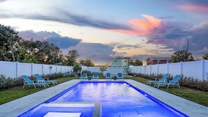 Une piscine rectangulaire dont le bord est illuminé de lumière bleue, entourée d’une clôture blanche et de chaises longues vertes.