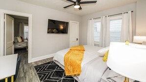 Et hotelværelse med seng, ventilator, fjernsyn, badeværelse med spejl og vindue med gardiner.