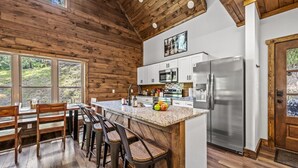 A kitchen with wooden walls, a large island with bar stools, stainless steel appliances, and a view through large windows.